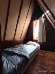 Interior kamar tidur kayu segitiga dengan jendela besar dan tirai putih.